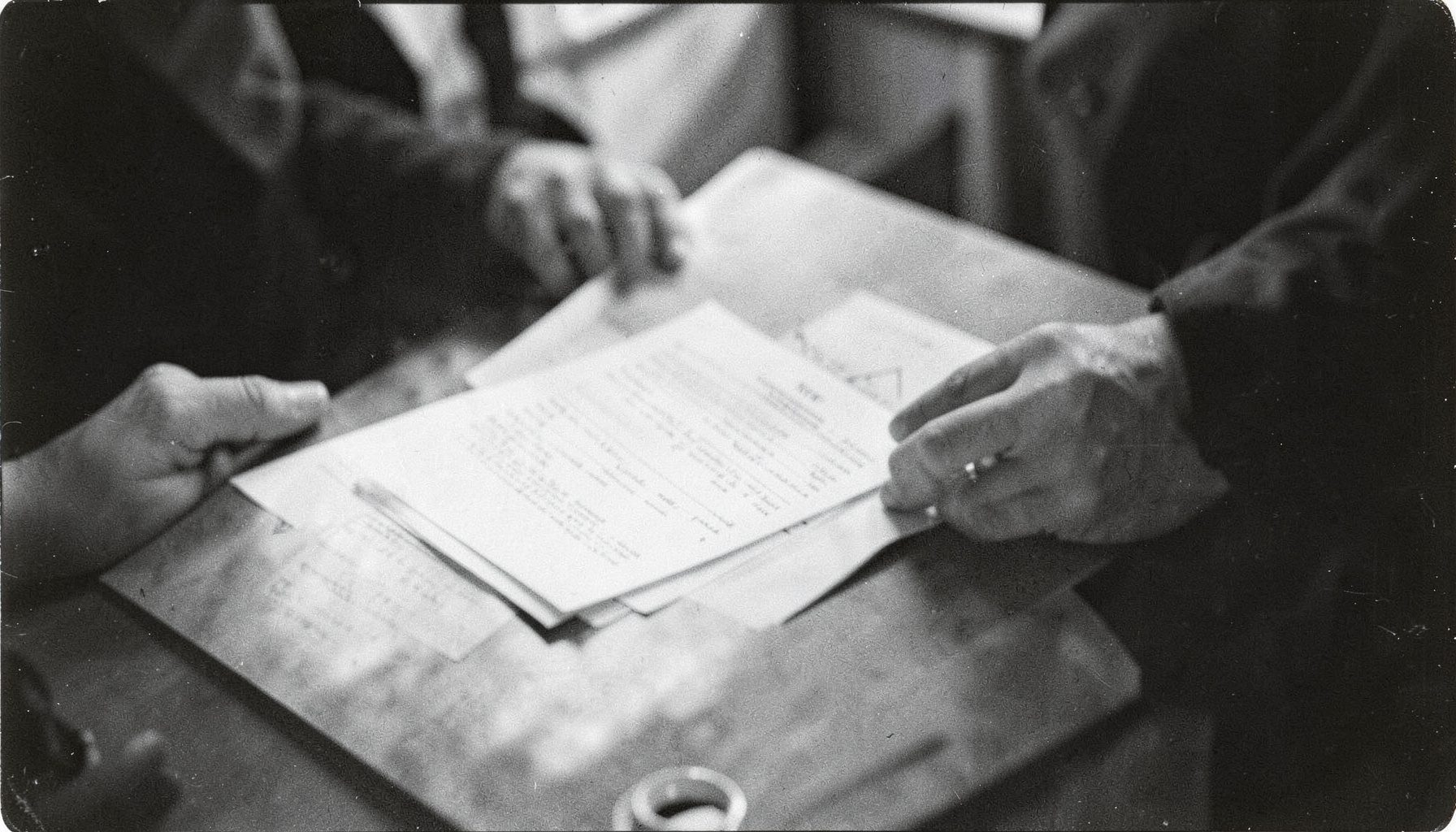 Fotografía analógica en blanco y negro de manos sosteniendo documentos sobre una mesa, con grano visible y atmósfera serena